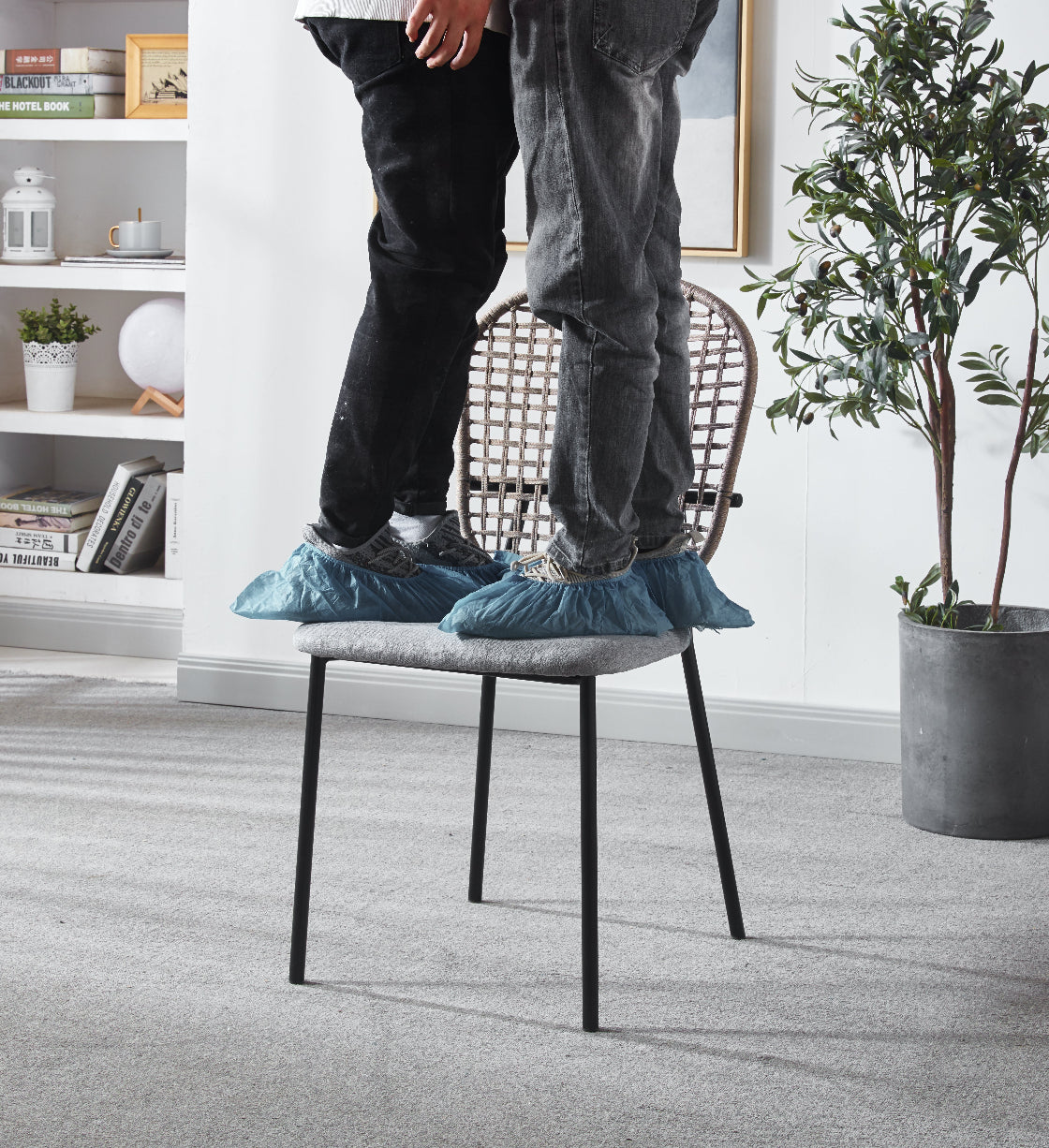 Two people using shoe covers on a chair in a room with plants and shelves.