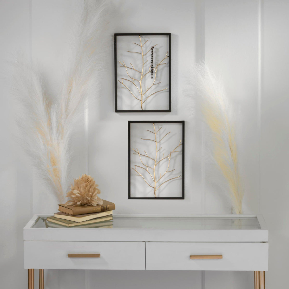 Two black-framed botanical wall art prints with gold-toned branches hanging above a white console table with gold handles, styled with books, a dried flower, and pampas grass against a white paneled wall.