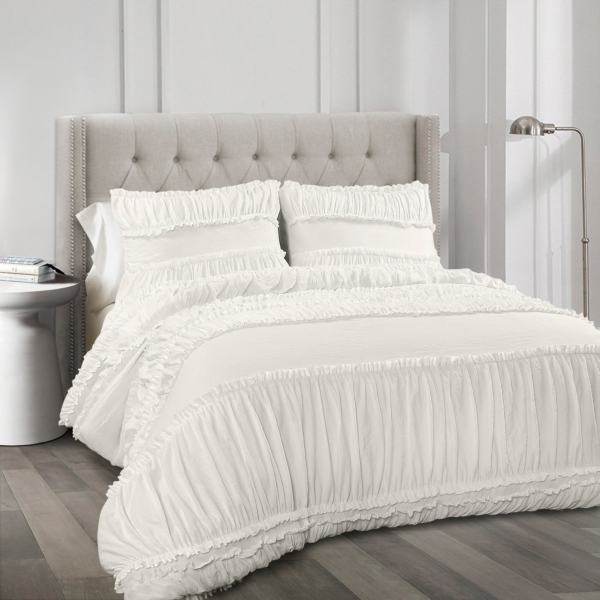White ruffled bedding set on a bed with a gray upholstered headboard.