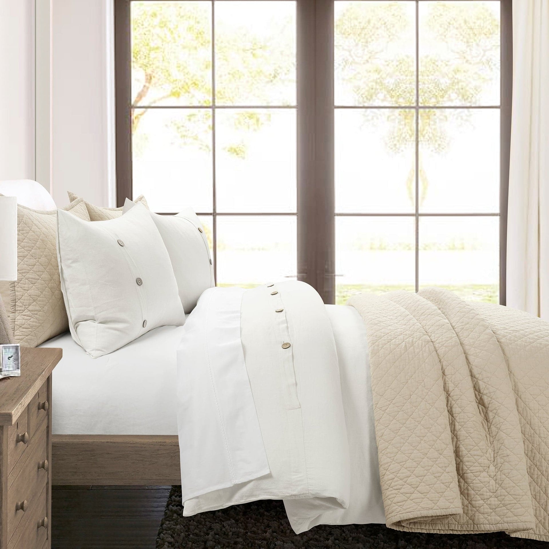 Neatly made bed with white bedding and beige quilt in a bright room with large windows.