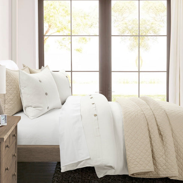 Neatly made bed with white bedding and beige quilt in a bright room with large windows.