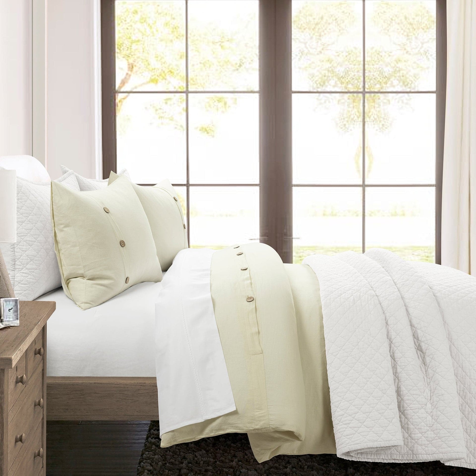 Neatly made bed with white bedding and pillows in a bright room with large windows.