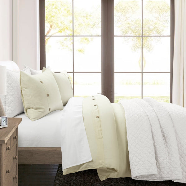 Neatly made bed with white bedding and pillows in a bright room with large windows.