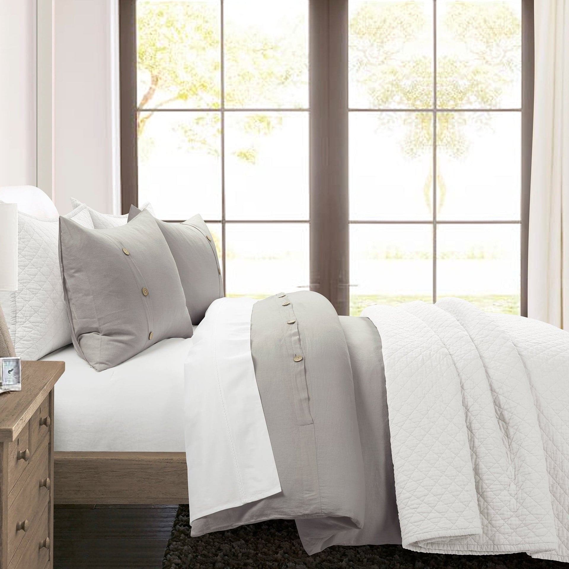 Neatly made bed with light-colored bedding in a bright room with large windows.