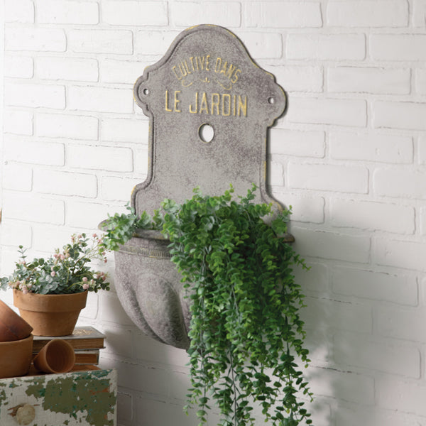 A metal wall water fountain planter with a sign that reads 'Jardin', mounted on a white wall with green plants spilling out from the fountain.
