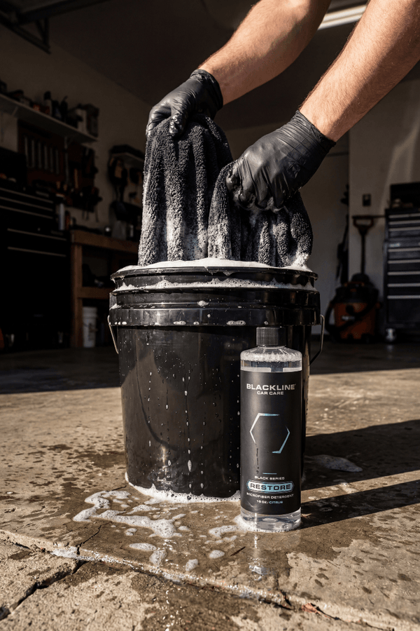 Person cleaning a black bucket with a sponge in a garage setting, with a bottle of Blackline Products Phoslock in the foreground.