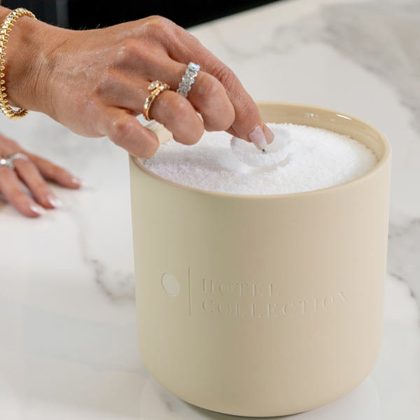 Hand reaching into a beige container of Hotel Collection bath salts on a marble surface.