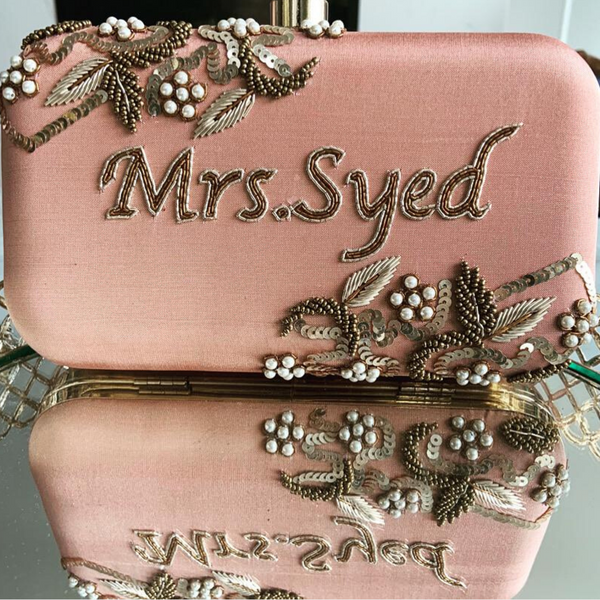 Pink clutch bag with 'Mrs. Syed' embroidery and decorative elements.