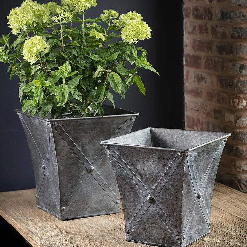 Two metallic planters with a plant on a wooden surface against a brick wall.