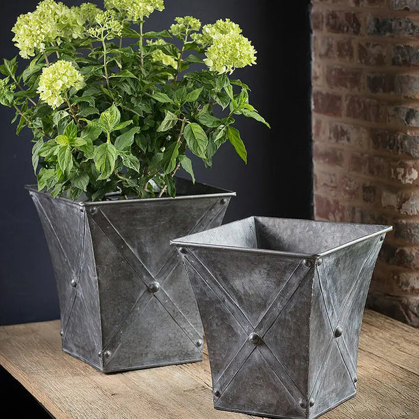 Two metallic planters with a plant on a wooden surface against a brick wall.