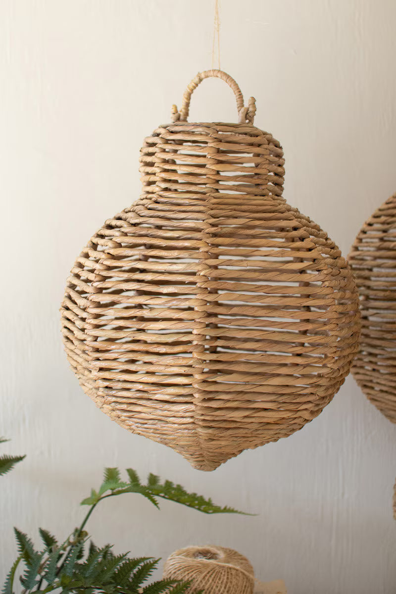 Woven rattan pendant light hanging against a neutral wall with a plant in the foreground.