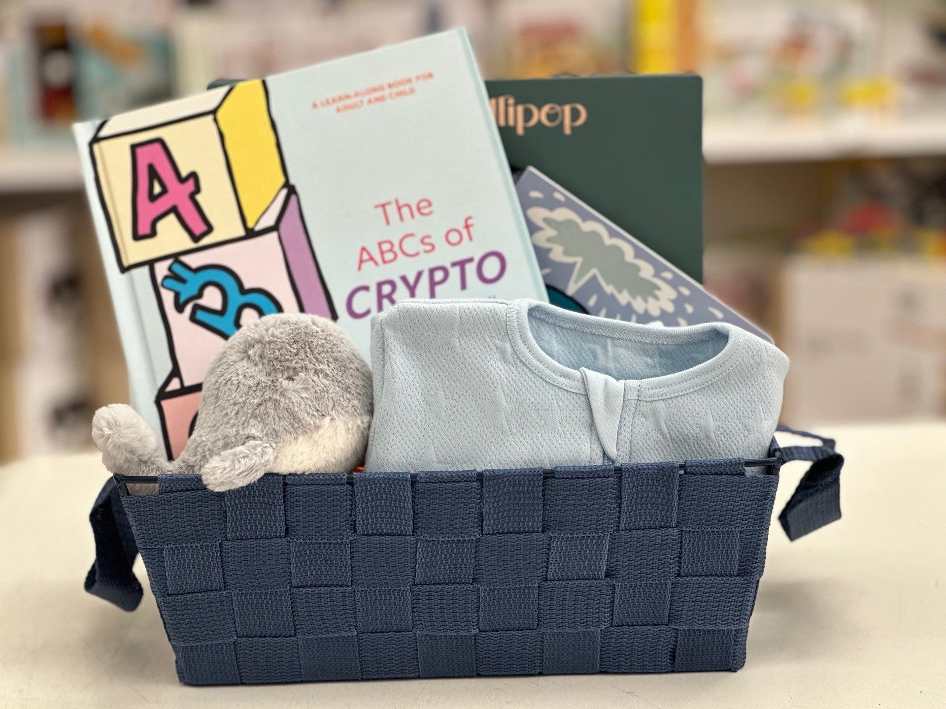 Navy blue basket with baby items including a book titled 'The ABCs of Crypto', a onesie, and a plush toy on a blurred background.
