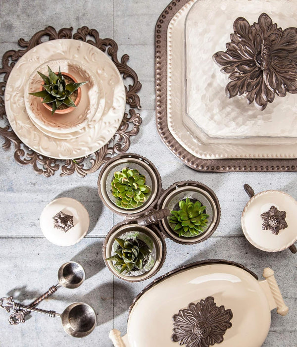 Acanthus Stoneware Pastry Keeper. Decorative table setting with ornate plates, bowls, and small potted plants on a textured surface.