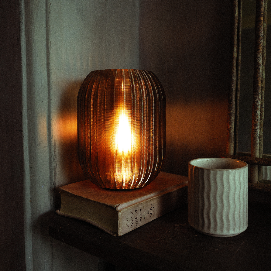 Wooden lamp with a warm glow on a shelf next to a white ceramic cup.