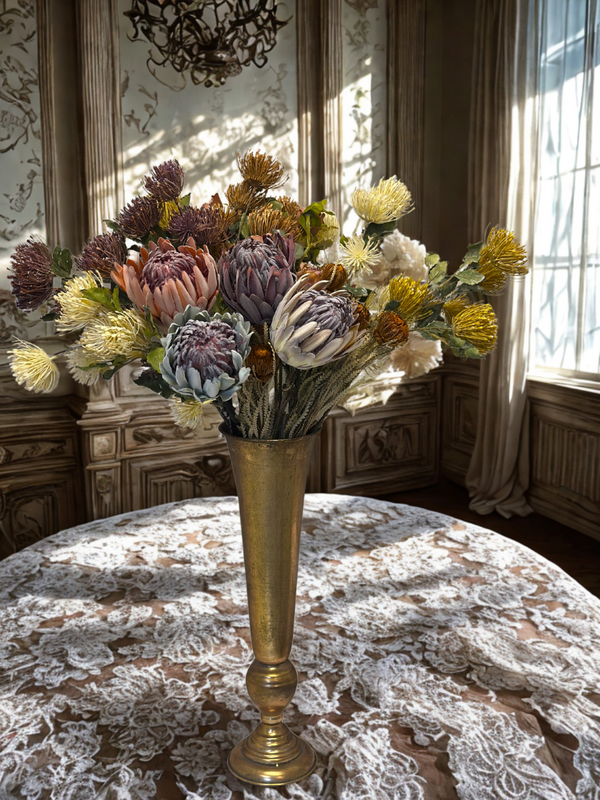Decorative floral arrangement in a gold vase on a lace tablecloth with sunlit windows in the background.