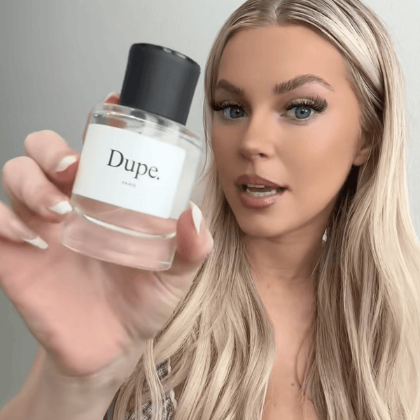 Woman holding a perfume bottle labeled 'Dupe' against a neutral background