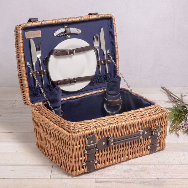 Open wicker picnic basket with cutlery, plates, and glasses on a wooden surface.