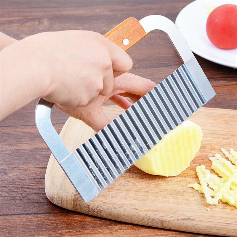 A hand holding a silver crinkle cutter with a wooden handle, cutting a waffle fry on a wooden board.