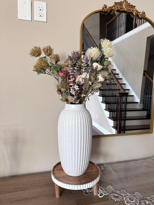 White vase with dried flowers on a wooden stand in front of a mirror reflecting a staircase.