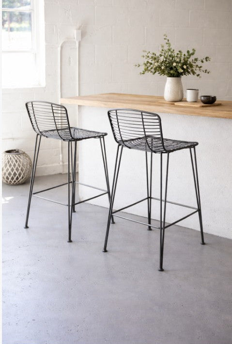 Kalalou Set of 2 - METAL BAR STOOL CHW1008. Two black metal stools in front of a wooden table with a vase of flowers on a light gray floor.