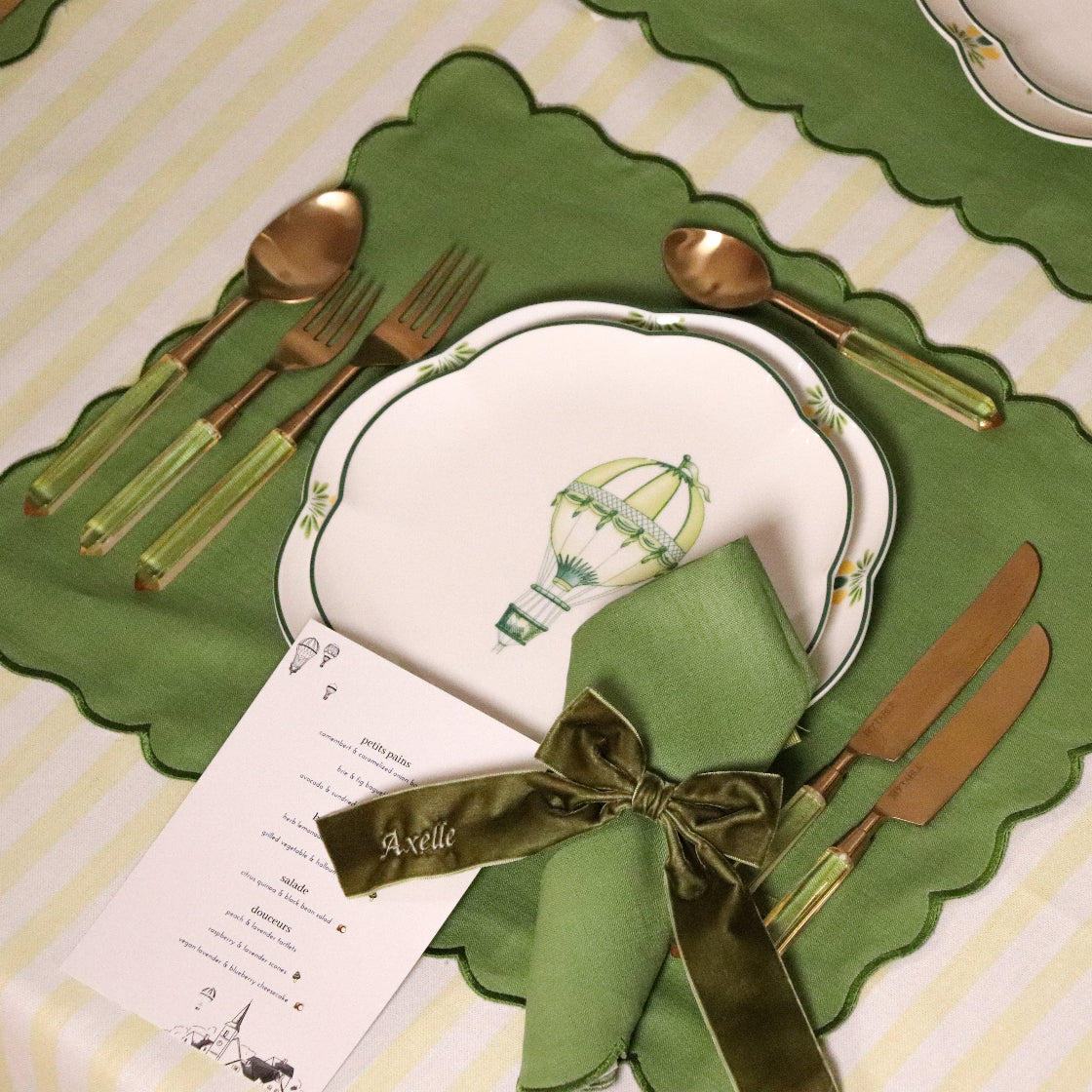 Dining table setting with green placemats, cutlery, and a napkin with a bow on a striped tablecloth.
