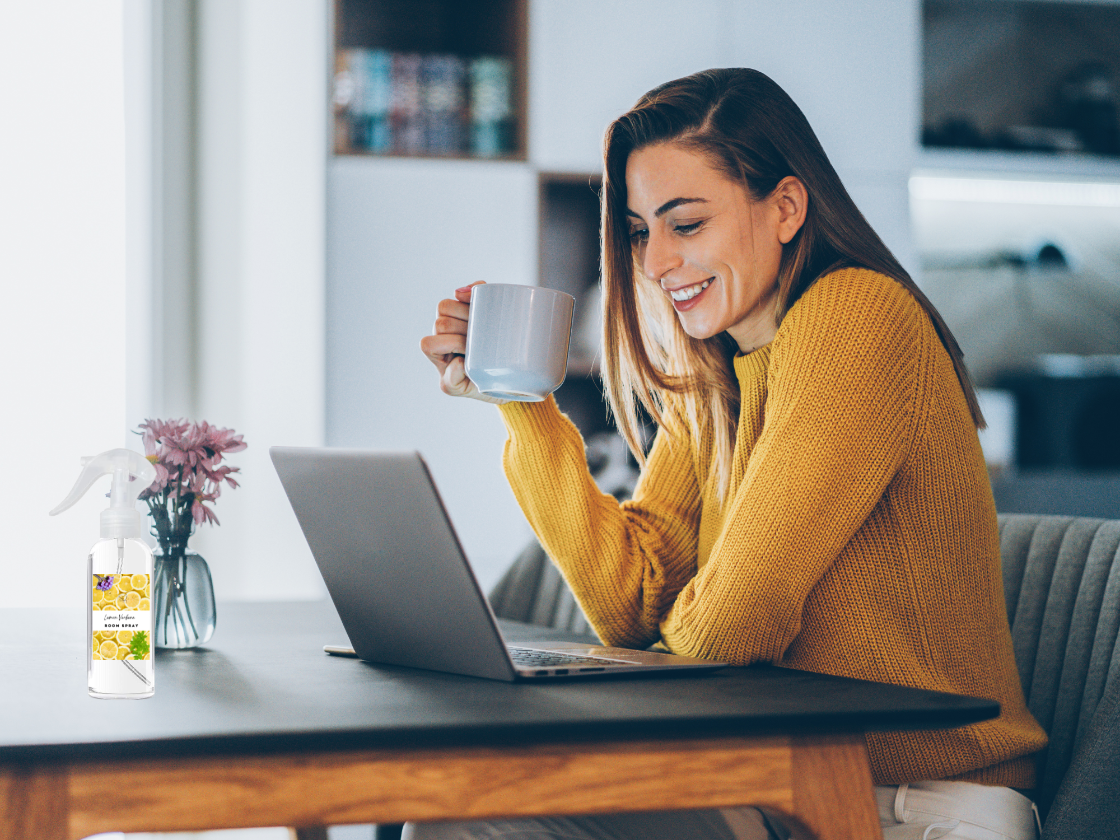 Woman in yellow sweater holding a mug and using a laptop with a bottle of cleaning spray on a table.