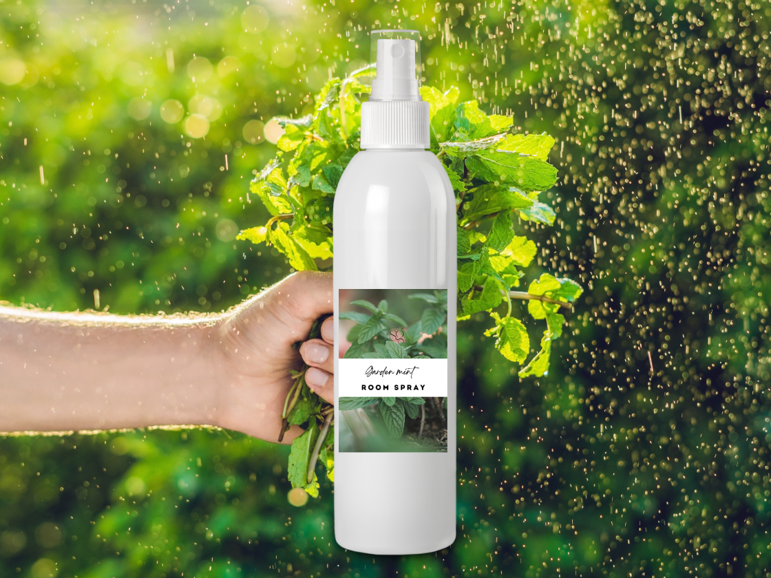 Hand holding a white room spray bottle labeled 'Jasmine Mint' with greenery in the background
