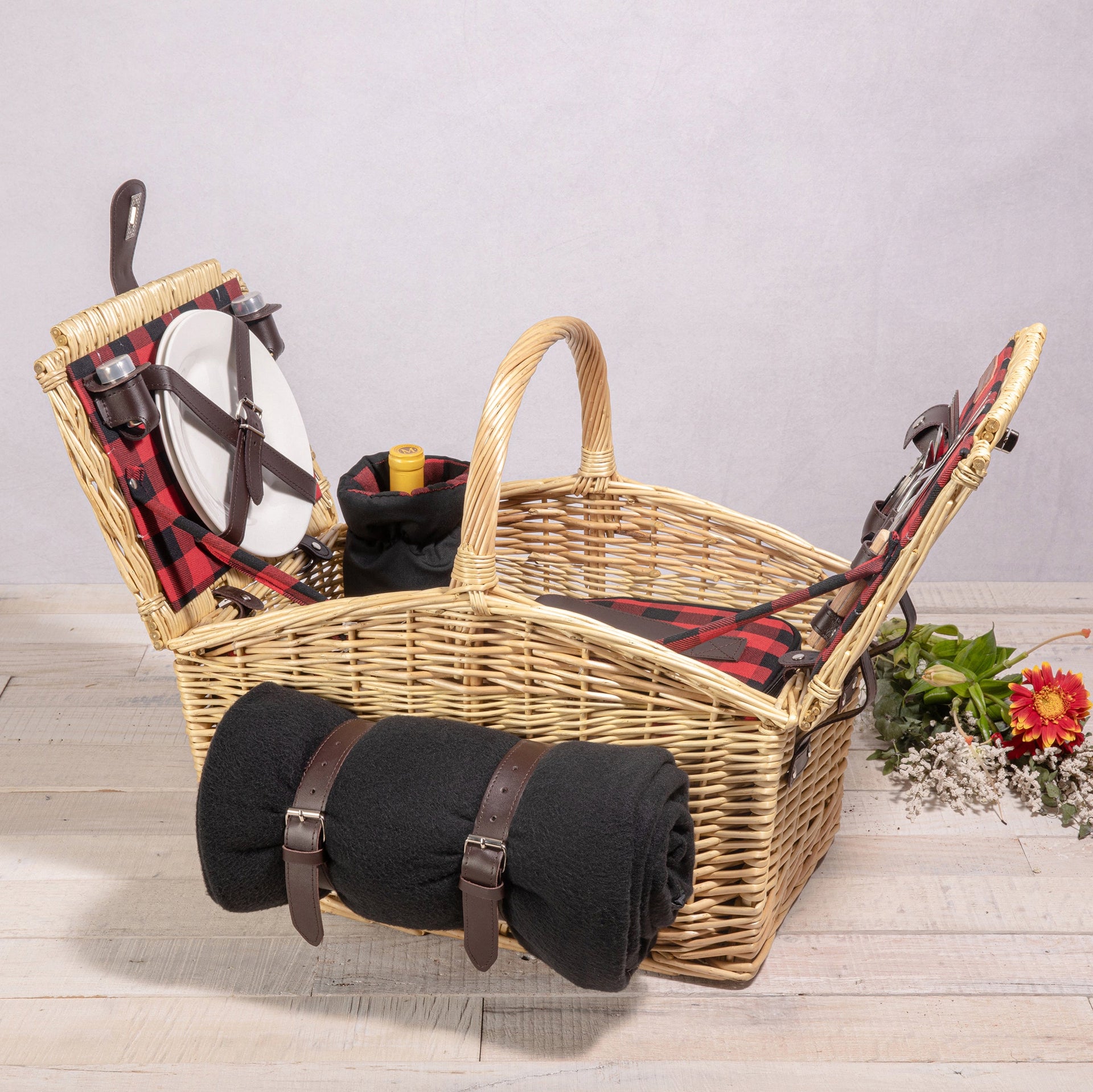 Wicker picnic basket with black blanket, glasses, and a bottle on a wooden surface.