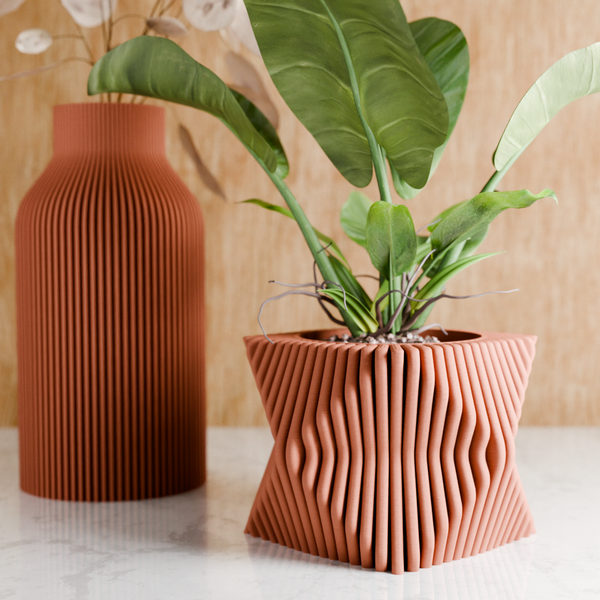 Decorative terracotta vase with wavy design next to a plant on a light surface.