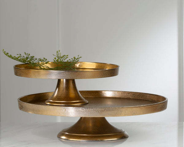 A two-tiered hammered brass cake stand or pedestal tray, styled with a small green sprig on the top tier, set against a clean white background.