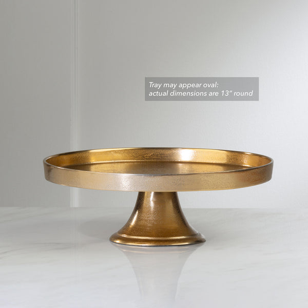 A round pedestal cake stand in a textured metallic gold finish, shown on a light gray background with a text overlay noting it may appear oval due to perspective.