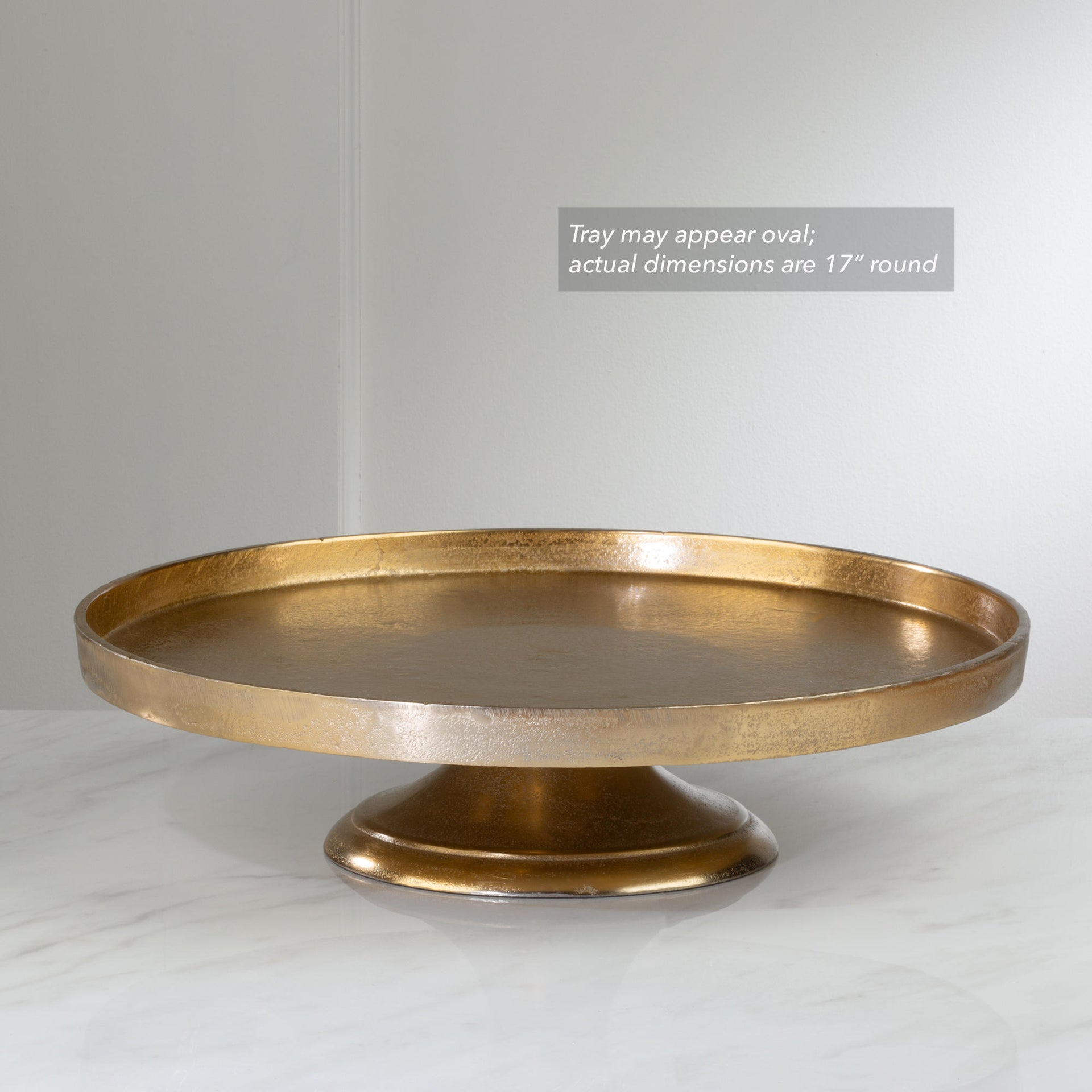 A round pedestal tray in a distressed gold finish, shown on a light marble surface against a neutral gray background. A text overlay in the upper right states 'Tray may appear oval; actual dimensions are 17