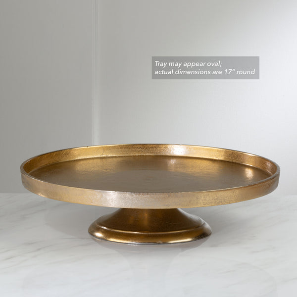 A round pedestal tray in a distressed gold finish, shown on a light marble surface against a neutral gray background. A text overlay in the upper right states 'Tray may appear oval; actual dimensions are 17