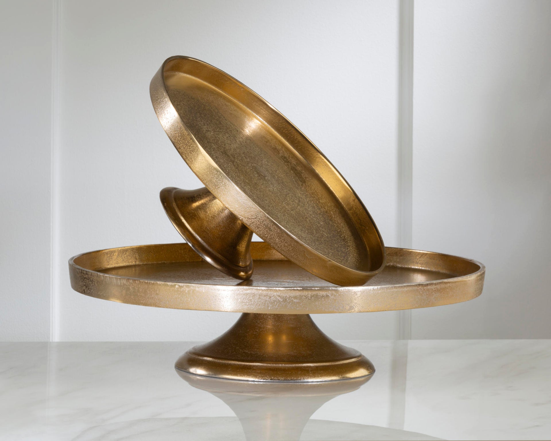 A decorative metallic pedestal stand with two round brass-toned plates, one flat circular tray resting on the base and another angled plate leaning against it, shown on a light gray studio background.