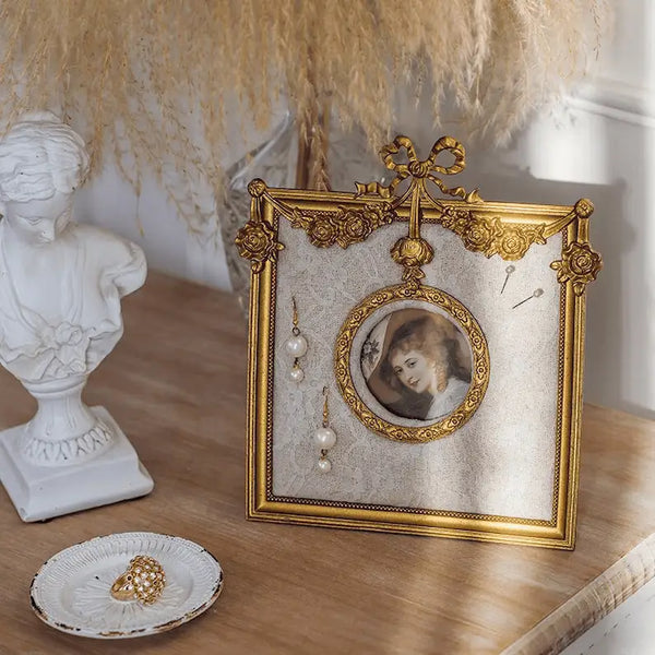 Decorative gold frame with a portrait and jewelry on a wooden surface with a white bust and decorative elements.