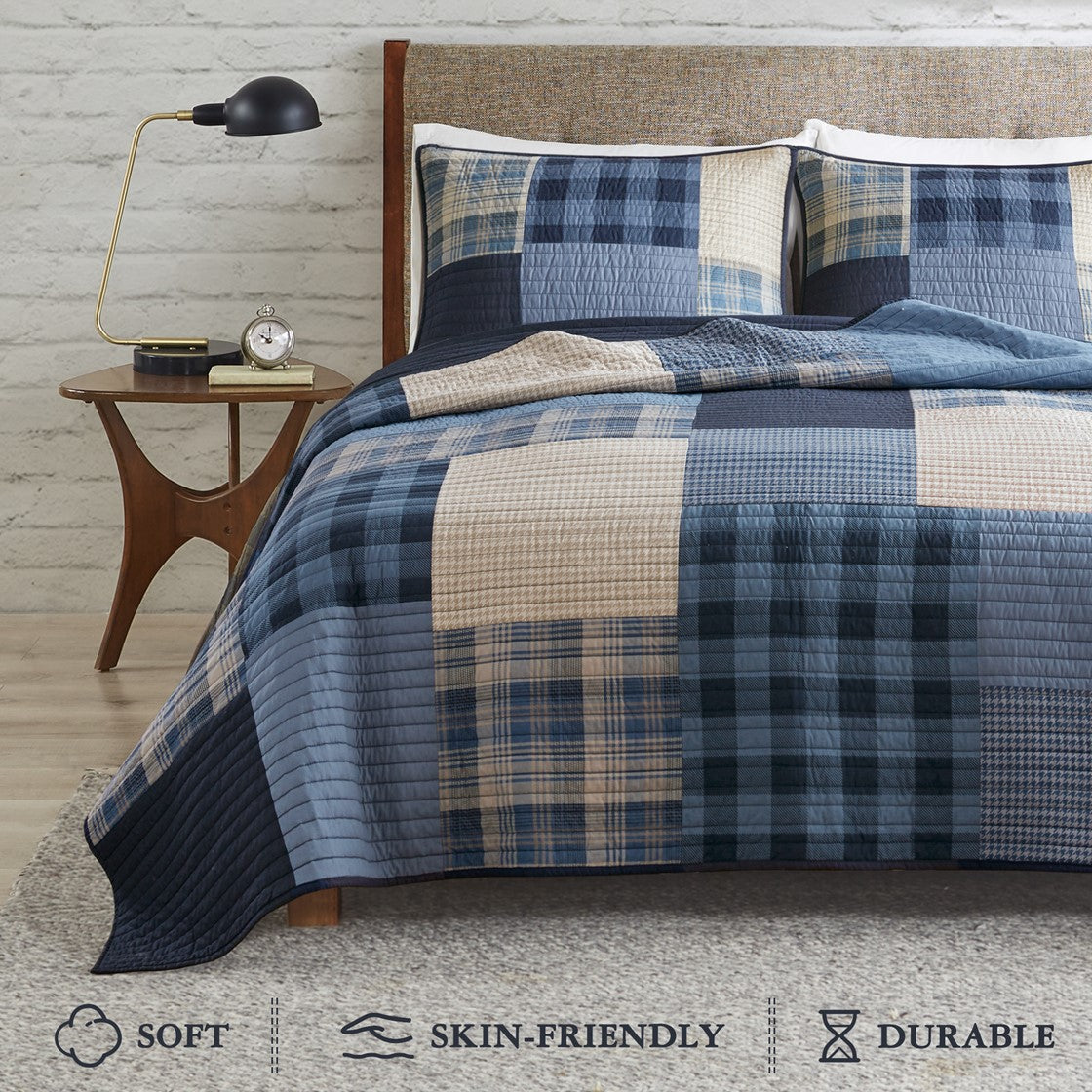 Winter Hills Oversized Cotton Quilt Set by Woolrich. Bed with blue and beige checkered quilt, nightstand with lamp and clock, wooden floor and gray wall.