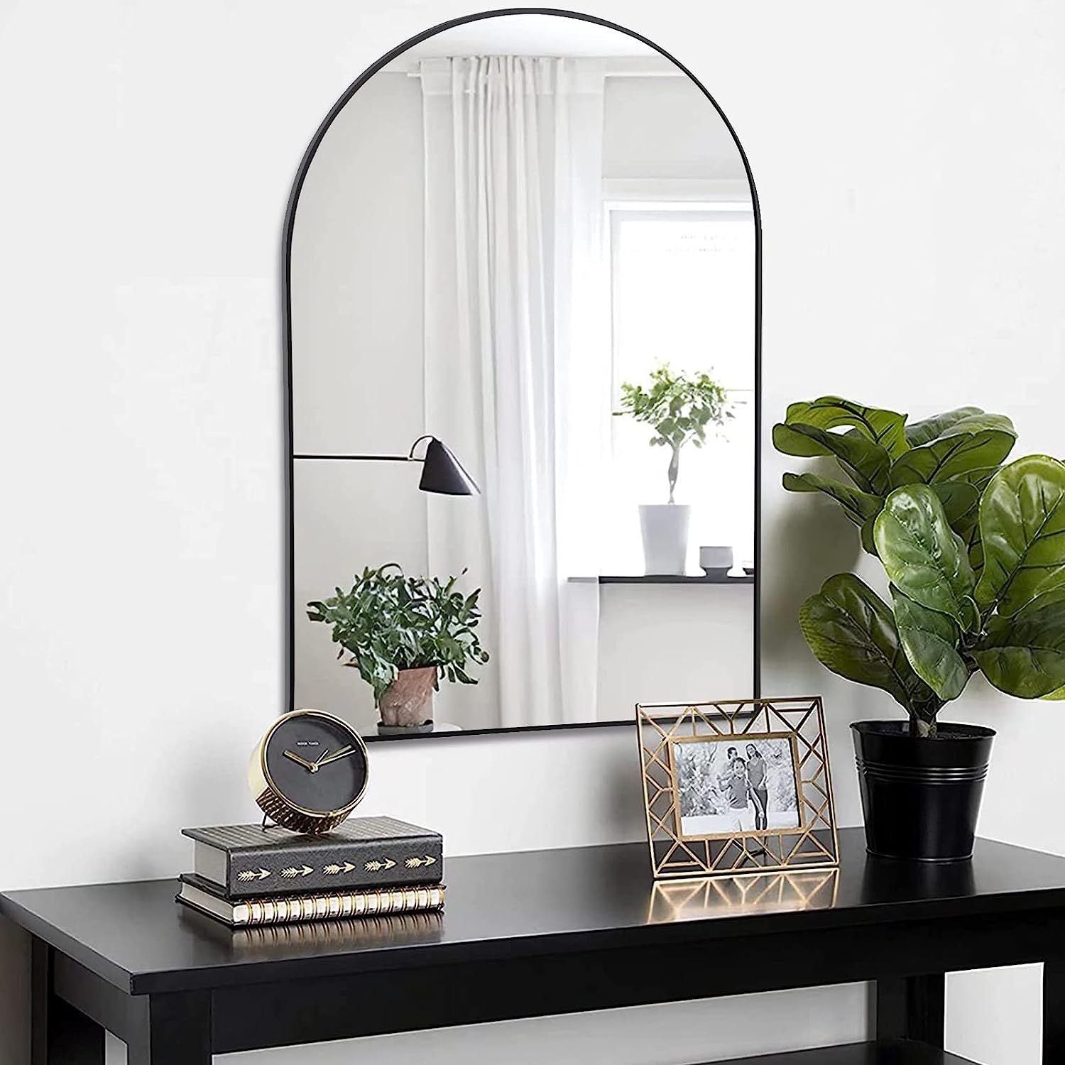 24x36 Inch Black Metal Framed Arched Wall Mirror hanging above a black desk with decorative items and greenery, enhancing a minimalist interior.