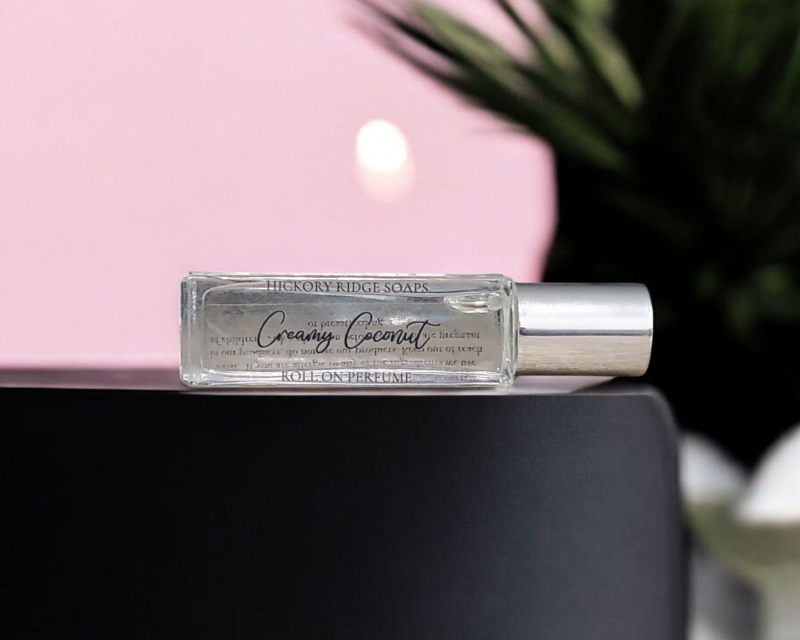 Roll-on perfume bottle labeled 'Creamy Coconut' by Hickory Ridge Soaps on a black surface with a pink background.