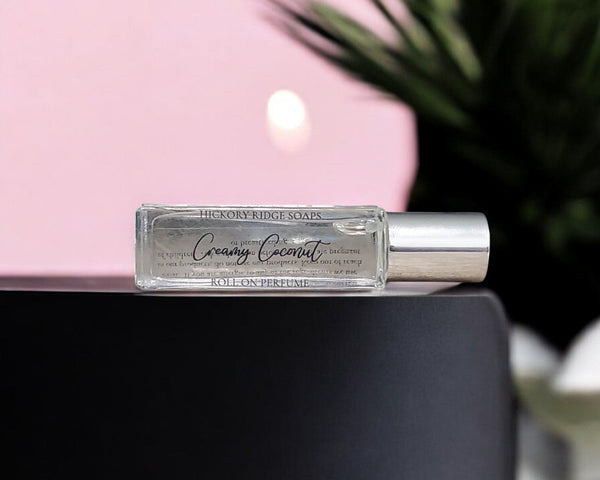 Roll-on perfume bottle labeled 'Creamy Coconut' by Hickory Ridge Soaps on a black surface with a pink background.