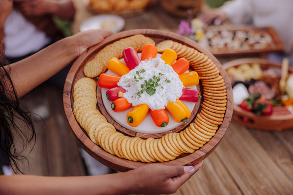 Person holding a round wooden board with crackers and a center dish, surrounded by blurred food items.