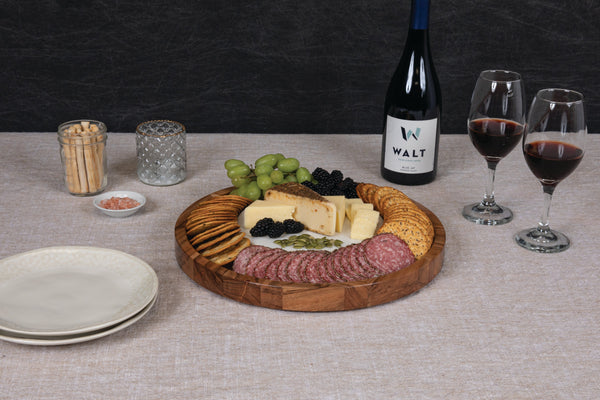 Wooden charcuterie board with meats, cheeses, and fruits on a table with wine and glasses.