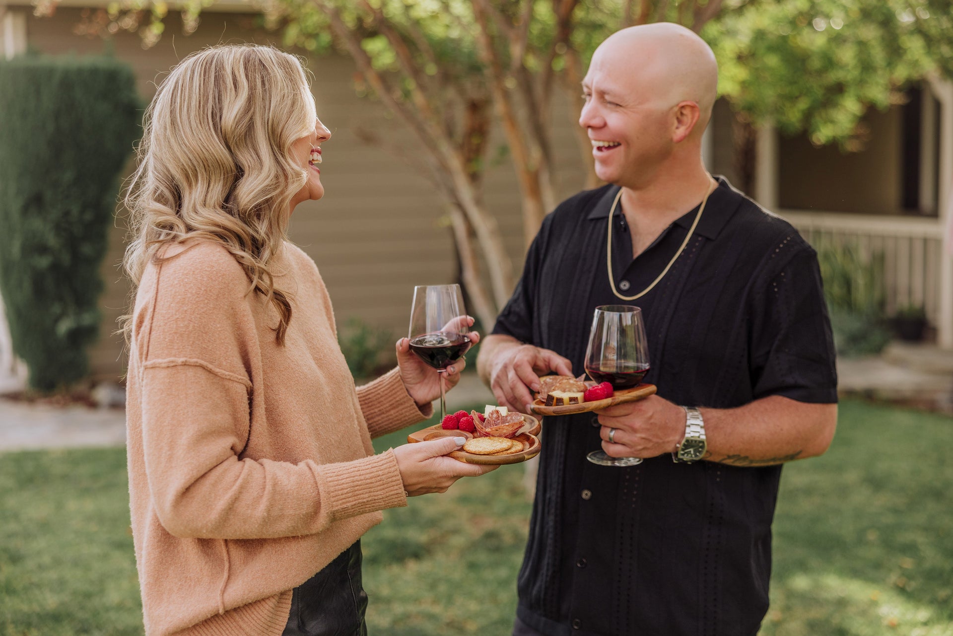 Two people standing outdoors, holding wine glasses and food, with a blurred background
