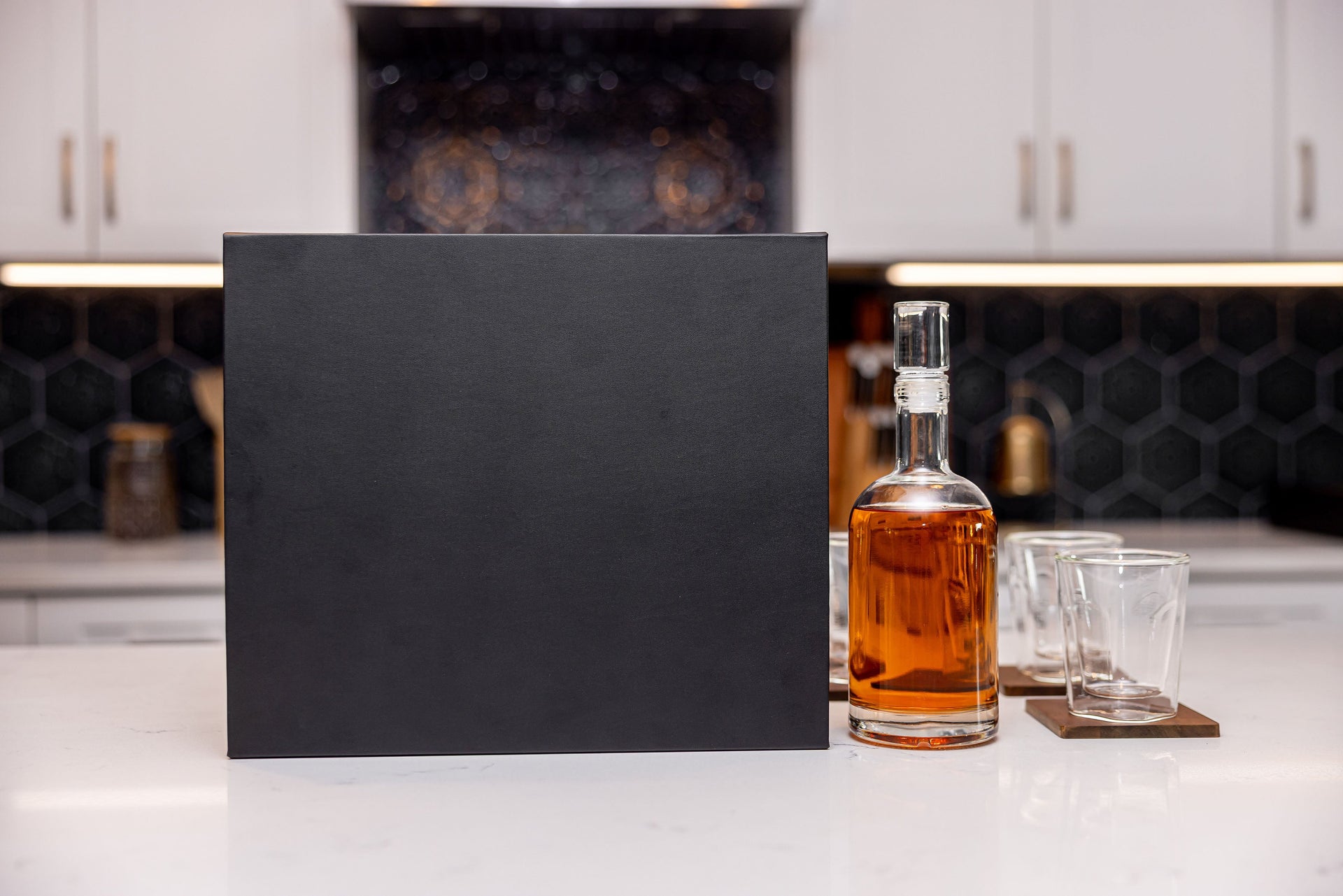 Black box on a kitchen counter with a bottle of amber liquid and two glasses.