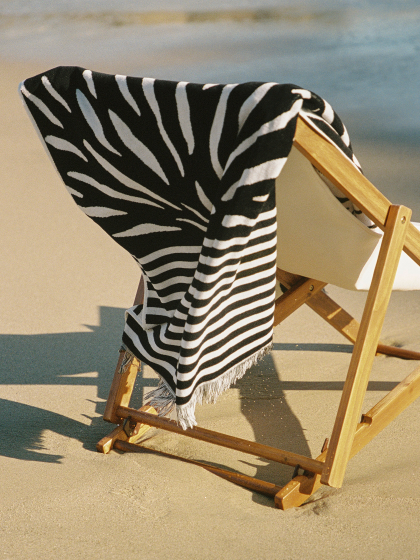 Zebra-patterned towel draped over a wooden chair on a sandy beach.