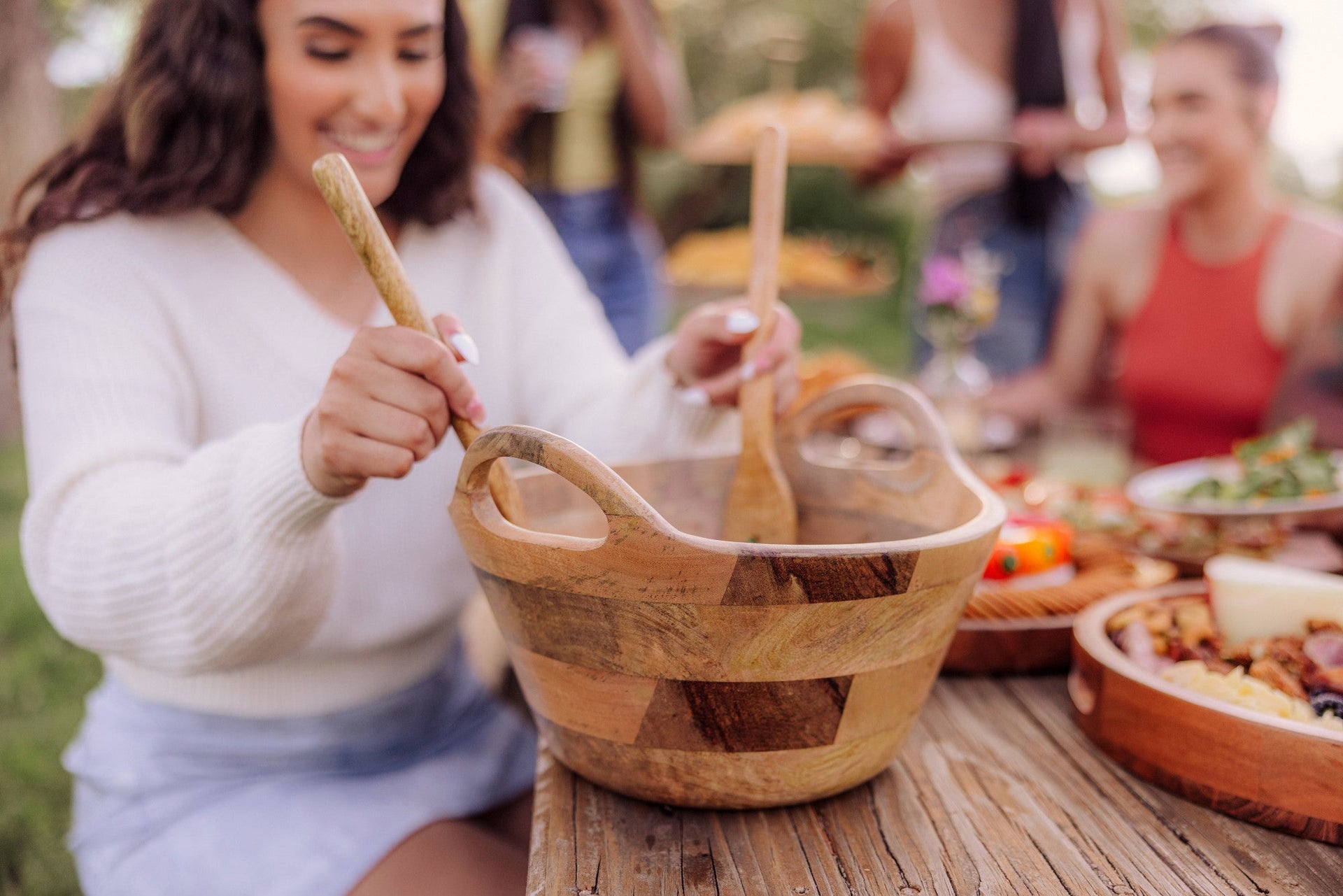 Woman stirring a wooden bowl at an outdoor gathering with food and people in the background.