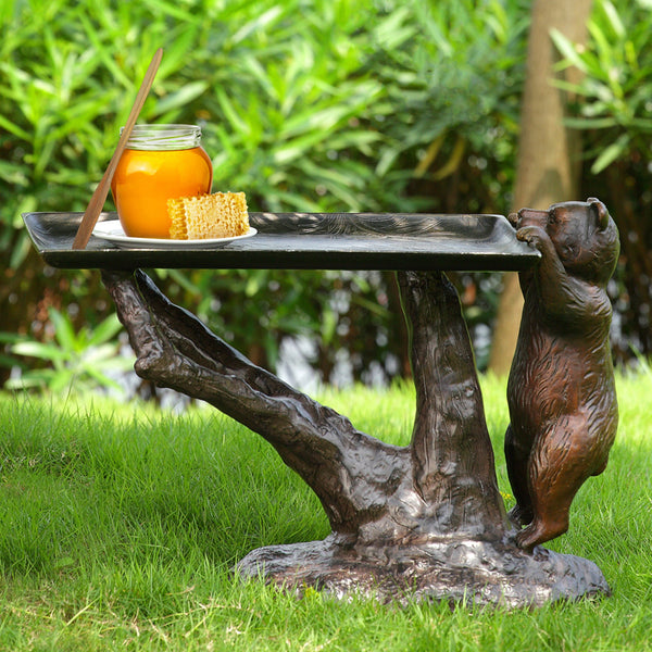 A bronze-colored bear-shaped accent table with a black top, featuring a jar of honey and some corn on top, set on a grassy area.