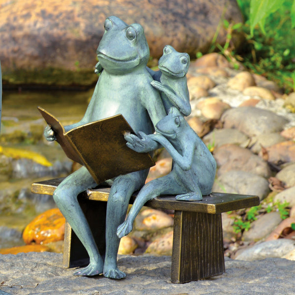 A bronze garden sculpture of a frog family with one frog reading a book to two smaller frogs, seated on a bench.